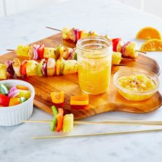 a wooden cutting board topped with skewers of fruit and veggies next to bowls of dipping sauce