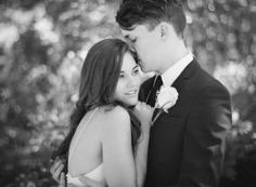 a man and woman standing next to each other in front of trees with their arms around each other