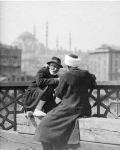 an old photo of two women sitting on a bench in front of a cityscape