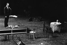 a man standing on top of a stage next to a table with chairs and a microphone