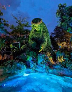 a large green monster statue sitting on top of a blue pool in front of trees