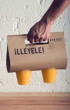 a person holding a paper bag with two yellow cups in front of it on top of a wooden table