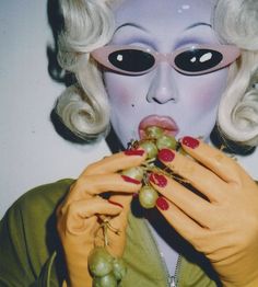 a woman with white hair and black eyes holding grapes in front of her face while wearing an alien mask