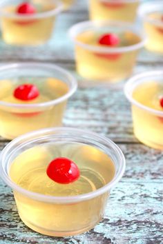 small plastic cups filled with yellow liquid and topped with a red cherry sitting on top of a wooden table