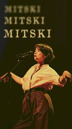 a woman standing in front of a microphone with her hands out to the side while wearing a white shirt and brown pants