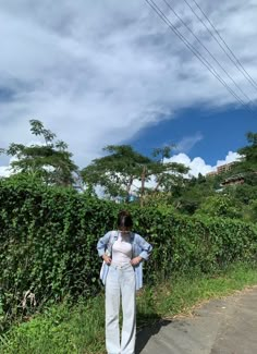 a woman standing on the side of a road next to a lush green field and bushes