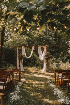 A beautiful garden wedding setup with a wooden arch draped in white fabric and adorned with floral arrangements, surrounded by string lights and wooden chairs under tall trees. Ceremony In The Woods, Nature Wedding Arch, Bohemian Vintage Wedding, Backyard Wedding Small Space, Small Wedding In The Mountains, Wedding Ideas Field, Wedding Alters Outside, Fall Wedding Trellis Ideas, Unique Wedding Altar Ideas