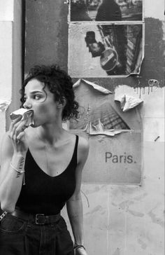 black and white photograph of a woman eating food in front of a wall with posters on it