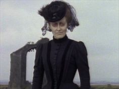 a woman in a black dress and hat standing next to a stonehenge with her hands on her hips