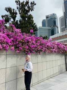 a woman standing next to a wall with purple flowers on it and buildings in the background