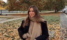 a woman standing in front of trees with leaves on the ground and wearing a scarf around her neck