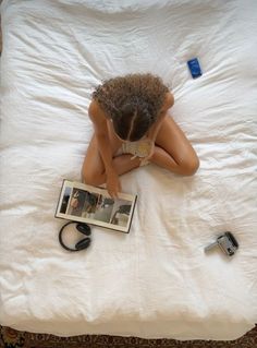 a woman laying on top of a white bed next to an open book and camera