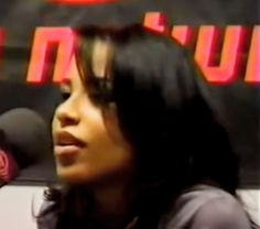 a woman talking into a microphone in front of a red and black sign with the word,