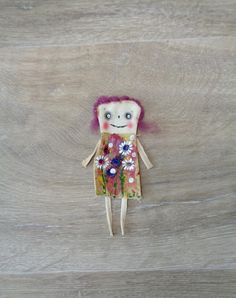 a small doll with pink hair and flowers on it's body sitting on a wooden surface