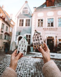 a person holding up a small model house in front of some buildings on a cobblestone street