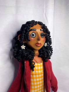 a doll with black hair wearing a red cardigan and yellow checkered dress is posed against a white backdrop