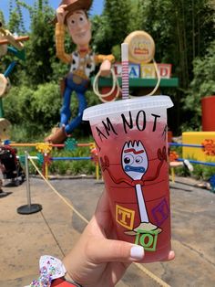 a person holding up a plastic cup in front of toy story land and the characters behind it