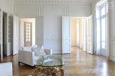 a living room filled with white furniture and wooden flooring next to two tall windows