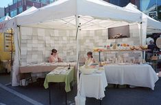 two people sitting at tables under a white tent with food on it's sides