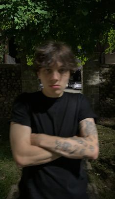 a young man with his arms crossed standing in front of a tree and building behind him