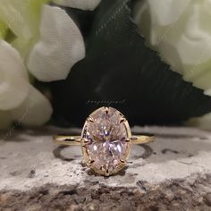 an oval shaped diamond ring sitting on top of a rock next to white flowers and greenery