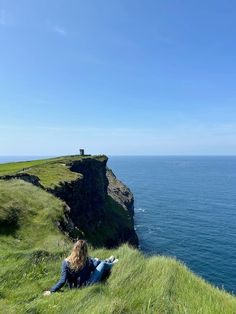 a woman sitting on top of a grass covered hill next to the ocean