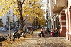 people are sitting at tables on the sidewalk in front of parked cars and trees with yellow leaves