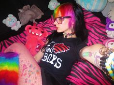 a woman with colorful hair and glasses laying on a bed next to many stuffed animals