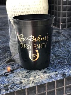 a black cup sitting on top of a counter next to a white vase with the name derby party written on it