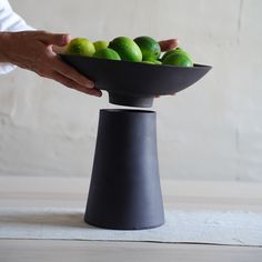 a person holding a black bowl filled with limes on top of a wooden table