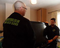 two men in black shirts are standing near a refrigerator