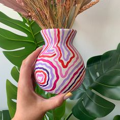 a hand holding a colorful vase next to a plant with long, thin stems in it