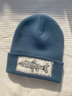 a blue beanie with a fish embroidered on the front and side, sitting on top of a bed