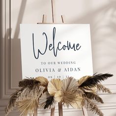 a welcome sign with feathers on it in front of a white wall and wooden easel