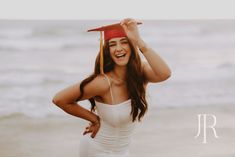 a woman wearing a graduation cap and gown on the beach with her hands behind her head