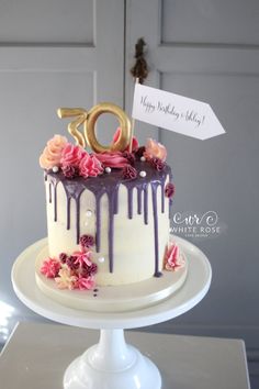 a white cake with purple icing and pink flowers on top, sitting on a pedestal