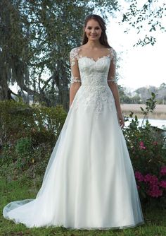 a woman in a white wedding dress standing on the grass near some trees and flowers