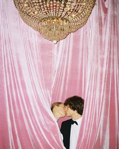 two people kissing each other in front of a pink curtain with a chandelier