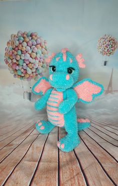 a blue stuffed dragon sitting on top of a wooden floor with balloons in the background