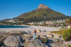 a woman standing on top of a rocky beach next to the ocean with a mountain in the background