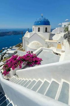 white buildings with blue domes and pink flowers on the steps leading up to them in front of water