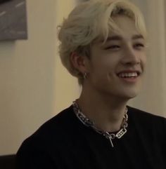 a young man with blonde hair smiling and wearing a black shirt, silver chain around his neck