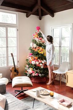 a woman standing next to a christmas tree in a living room with flowers on it