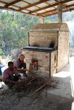 two men sitting in front of an outdoor oven with logs on the ground next to it