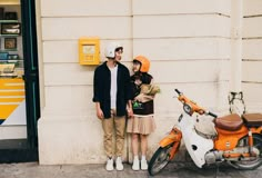 two people standing next to an orange scooter on the side of a building