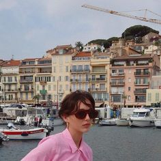 a woman wearing sunglasses standing in front of a harbor with boats and buildings behind her