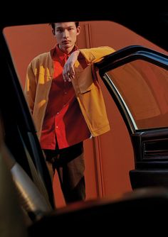 a man standing next to a car with his hand on the door handle and wearing a red shirt