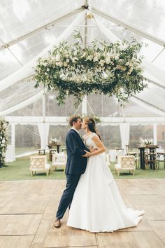 a bride and groom kissing in front of a white tent with flowers hanging from the ceiling