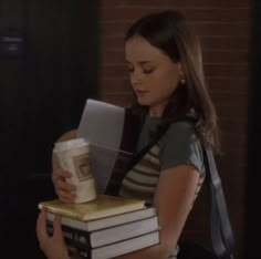 a woman holding two books and a cup in her hand while standing next to a brick wall