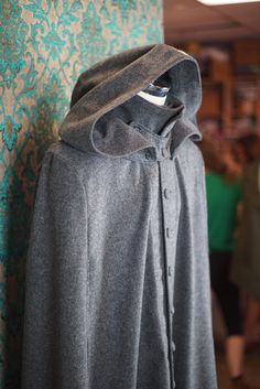 "ABOUT THIS STYLE: An incredible floor length cape that is both warm and fashionable. This style features a collar that can be worn either popped up with a detachable \"kidney\" throat latch or folded down. The detachable opera (or Jedi?) hood is double layered and very warm and water resistant. The piece does not have sleeves (unlike our caped coats) but instead has arm slits that you can stick your arms through. The cape is made from an ultra luxurious wool melton that is woven in England by H Winter Cape For Costume Events, Winter Costume Cape With Long Sleeves, Winter Long Sleeve Cape For Costume, Winter Costume Cape Outerwear, Winter Costume Cape, Fitted Fall Cape For Costume, Fitted Cape For Fall Costume, Fitted Winter Cape With Cape Sleeves, Fitted Winter Outerwear With Cape Sleeves